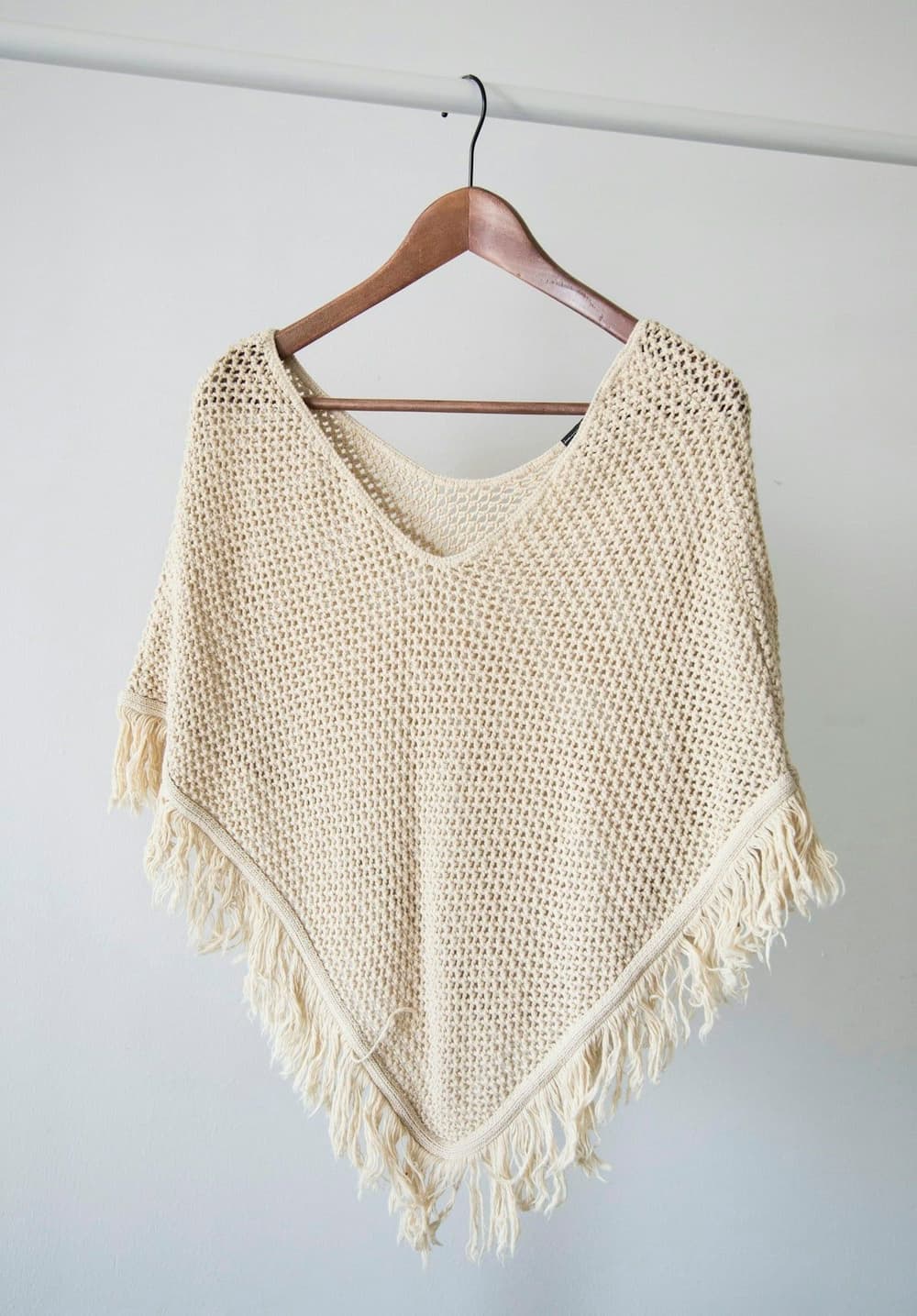 A hand-knit cream poncho hanging on a wooden hanger