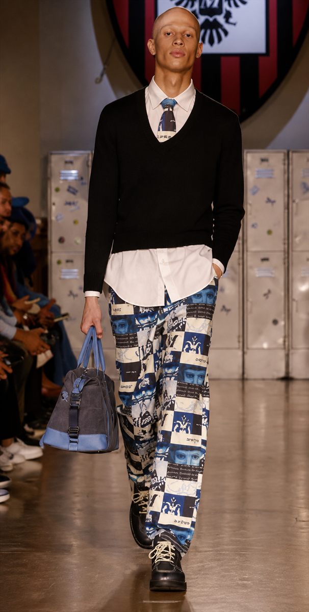 Model on the runway in a black V-neck sweater over a shirt and tie with printed trousers